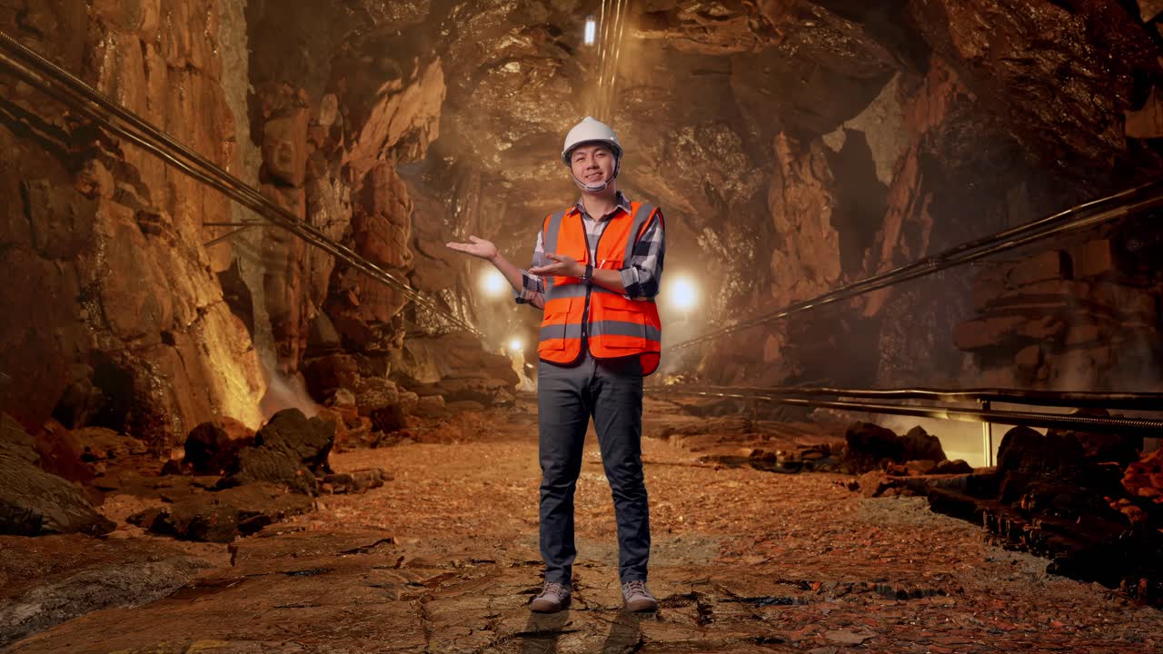 Full Body Of Asian Male Engineer With Safety Helmet Smiling And Pointing To Side While Standing In Underground Mine Tunnel