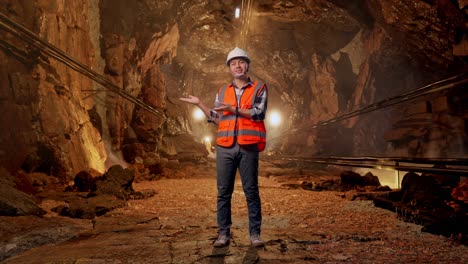 full body of asian male engineer with safety helmet smiling and pointing to side while standing in underground mine tunnel