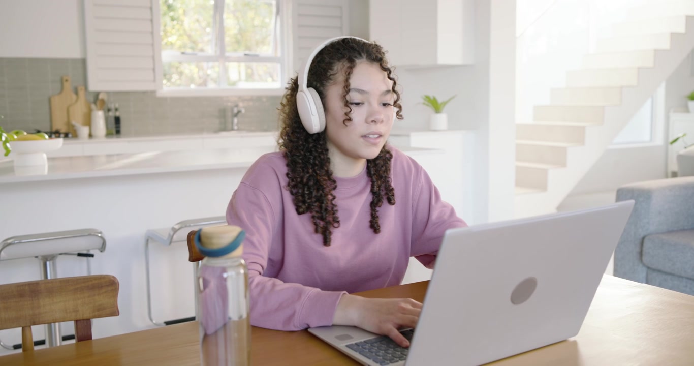 African American teen with headphones typing on laptop at kitchen table studying with water bottle