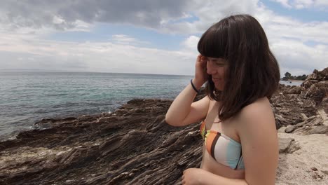 A-brunette-girl-in-her-20s,-wearing-swimwear-and-smiling-at-the-camera-on-a-rocky-coast-in-the-Mediterranean-on-a-partly-sunny-and-windy-day