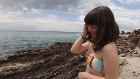 una chica morena de unos 20 años, vestida con traje de baño y sonriendo a la cámara en una costa rocosa en el mediterráneo en un día parcialmente soleado y ventoso