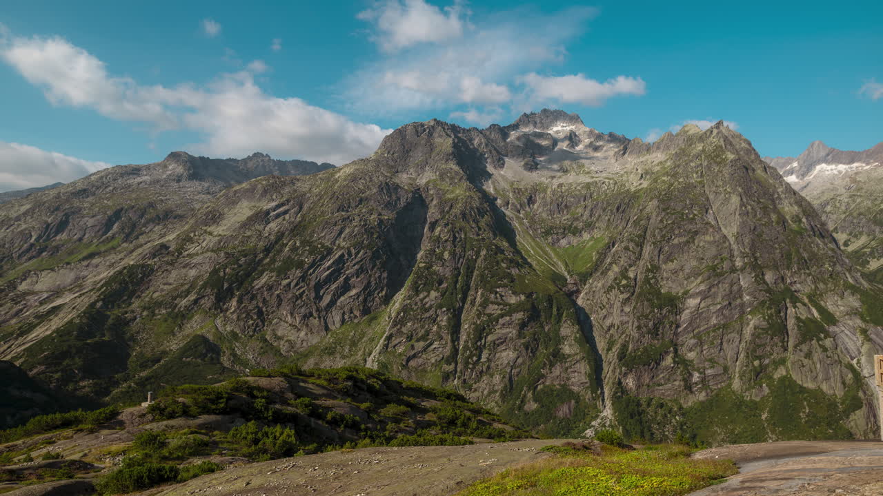Timelapse of rugged alpine mountains under a clear blue sky, with shifting clouds drifting across steep rocky peaks and green valleys