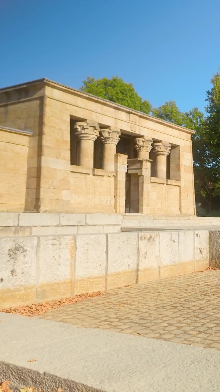 Una toma panorámica vertical del Templo de Debod, un antiguo templo egipcio reubicado en Madrid, España. Con arquitectura de piedra y exuberantes árboles verdes bajo un sol brillante.