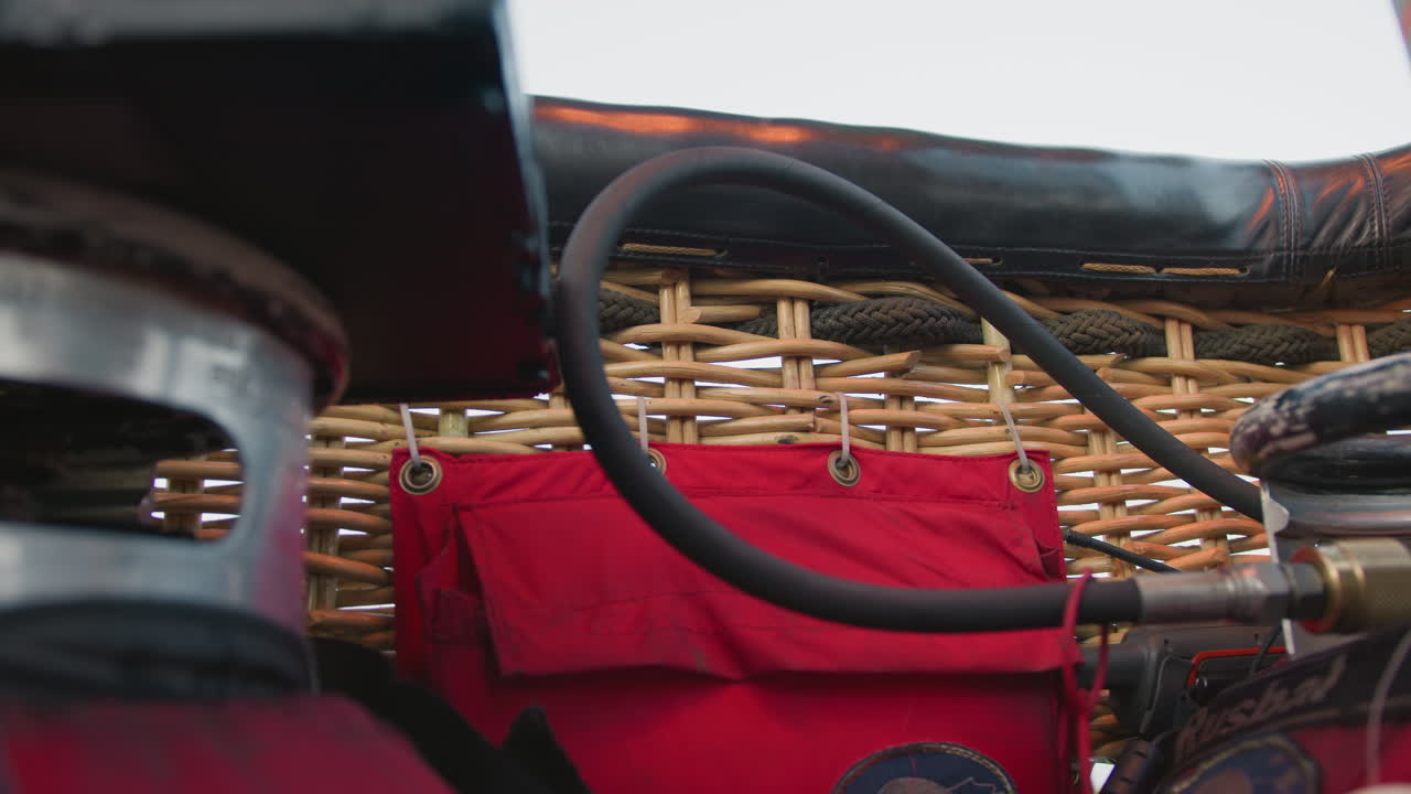 close up of hot air balloon burner system showing fuel hose connected to metal burner frame with wicker basket in background and red fabric storage pouch