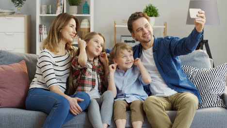 Alegre-Madre,-Padre,-Hija-E-Hijo,-Sentados-En-El-Sofá-De-La-Sala-De-Estar-Y-Divirtiéndose-Mientras-Hacen-Divertidos-Selfies-En-El-Smartphone,-Sonriendo-Y-Posando