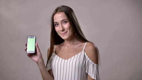 young excited girl is showing green screen of smartphone, watching at camera, smiling, communication concept, grey background