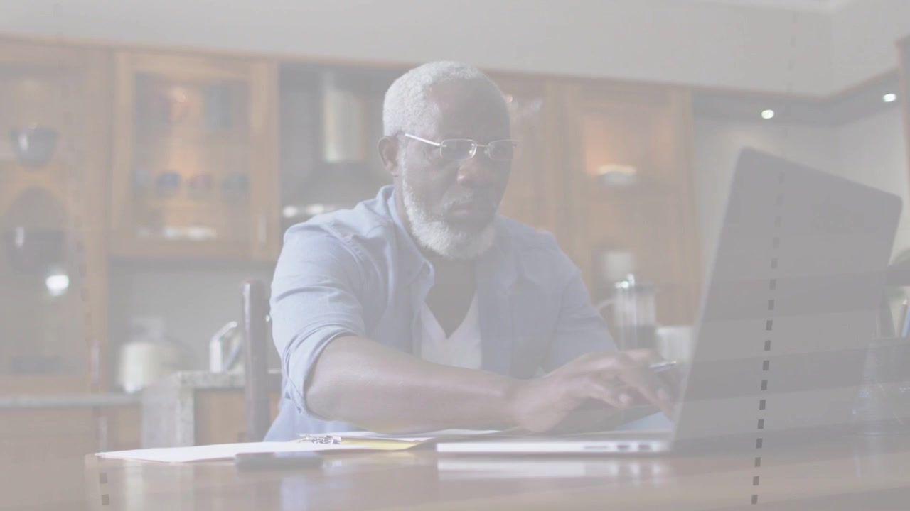 Senior man at kitchen typing on laptop adding dotted overlays checking business docs reaching phone