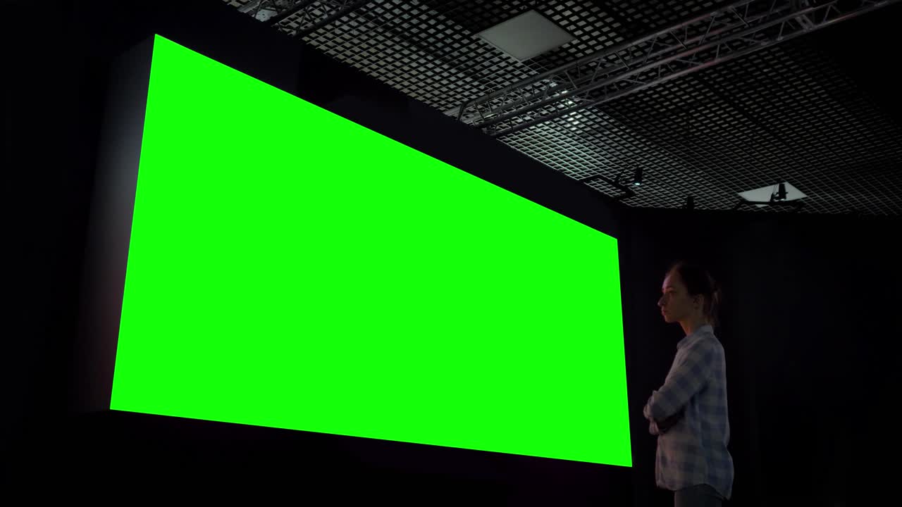 Woman looking at large blank green screen in dark room - chroma key concept