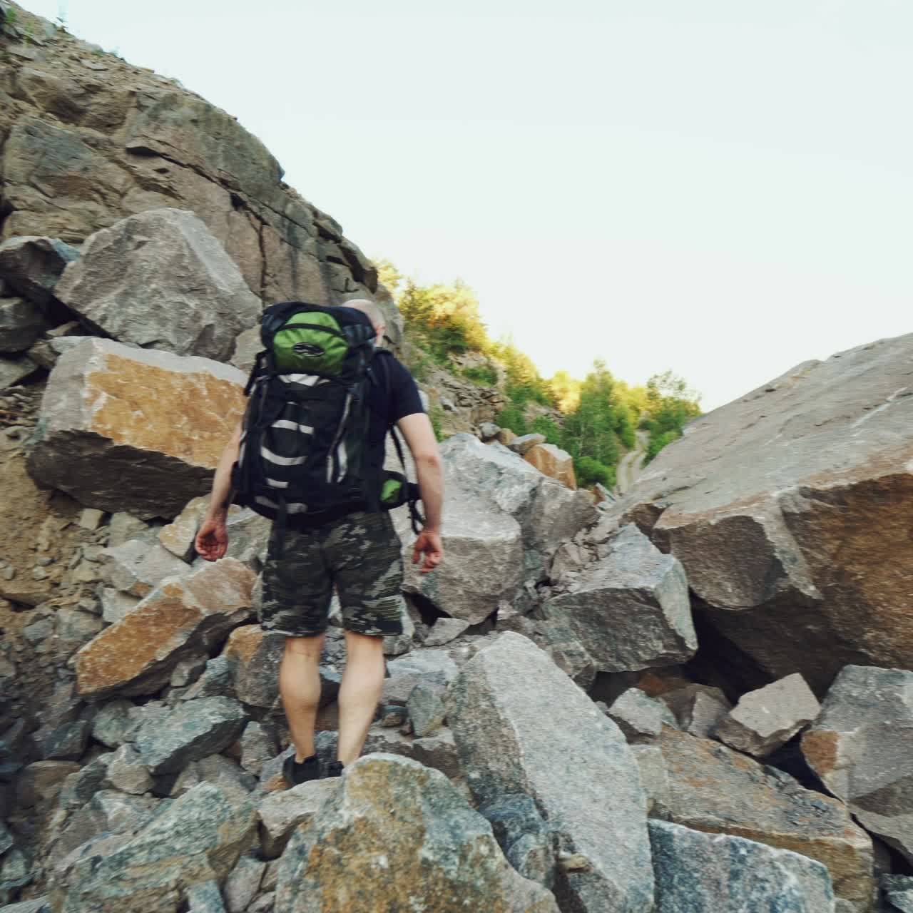 extreme tourist in a black t-shirt and shorts with a backpack on his shoulders is walking along the rocks and climbing a large stone in a warm weather in the summer