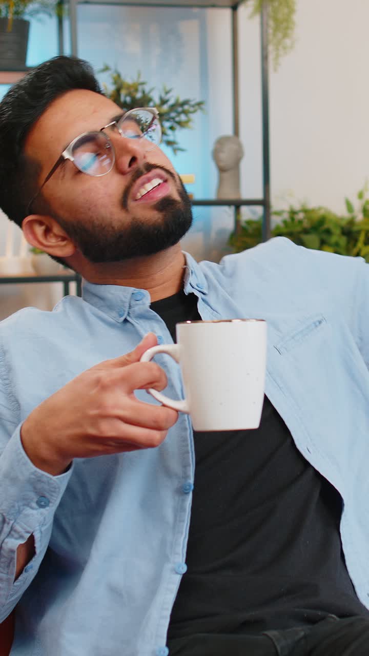 Træt, smilende indisk mand, der drikker en kop varm kaffe eller urtete om morgenen og slapper af.