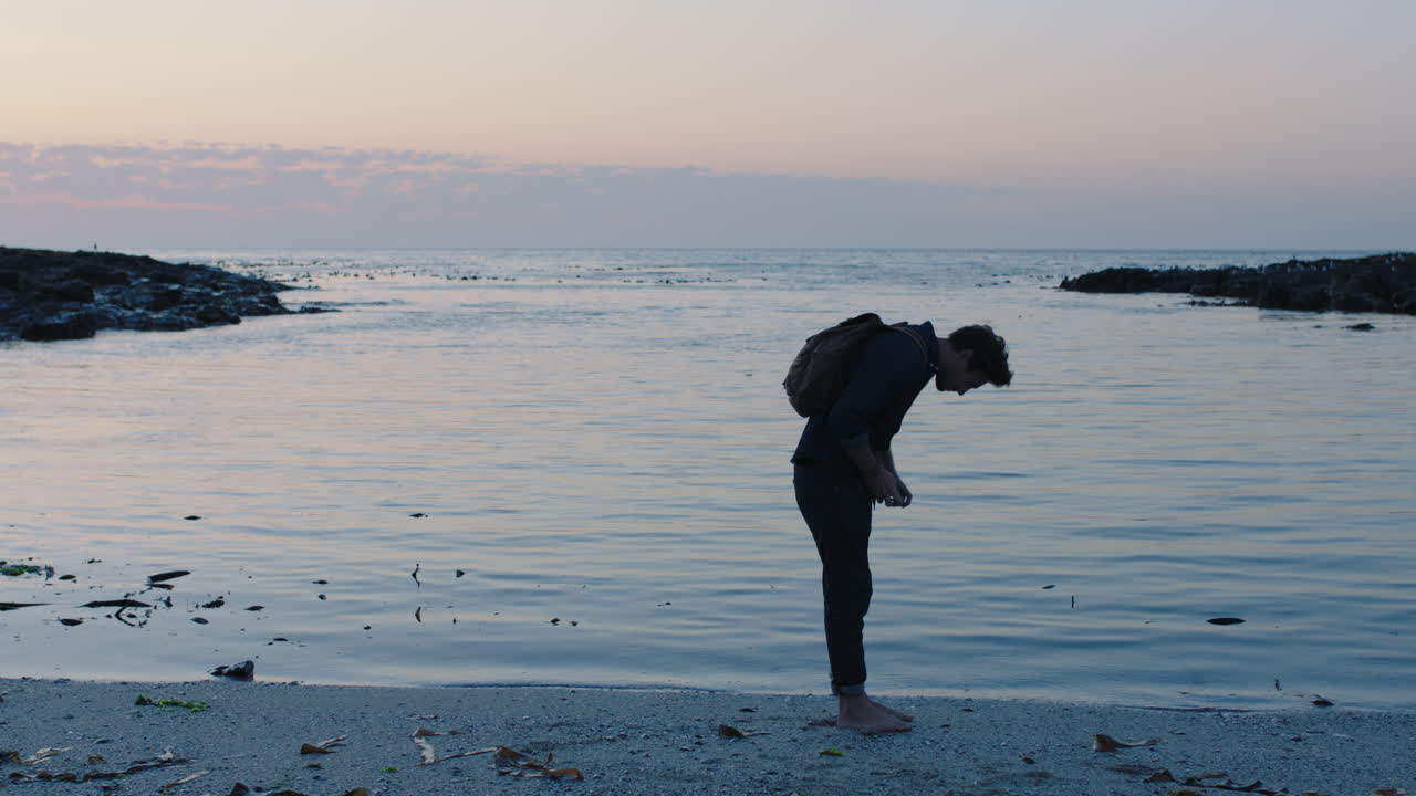 retrato de un joven turista caminando por la playa al atardecer usando un teléfono tomando fotos