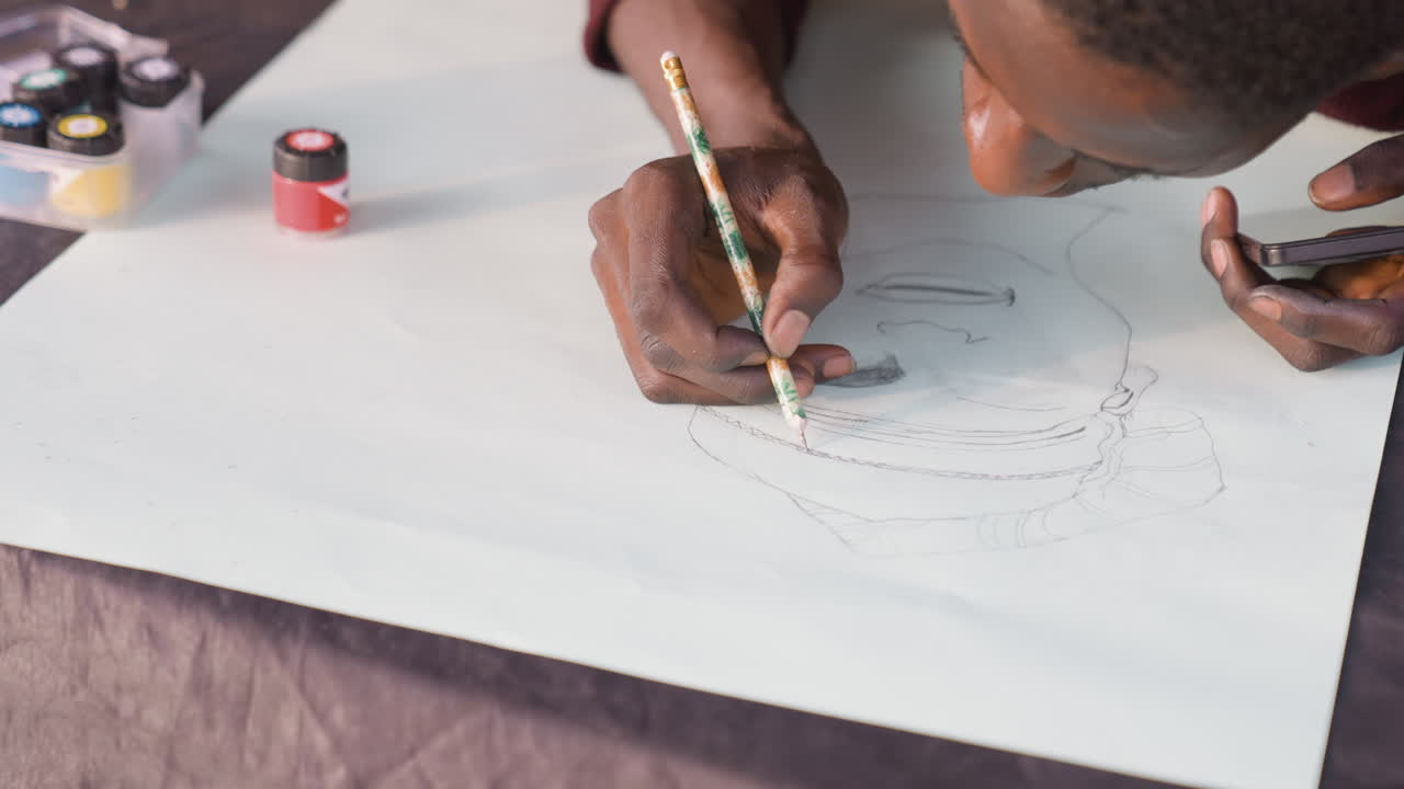 Close up hand painting black portrait. Intimate closeup of black male artist sketching delicate portrait lines with pencil on paper, jars of paint visible, calm studio mood and focused technique