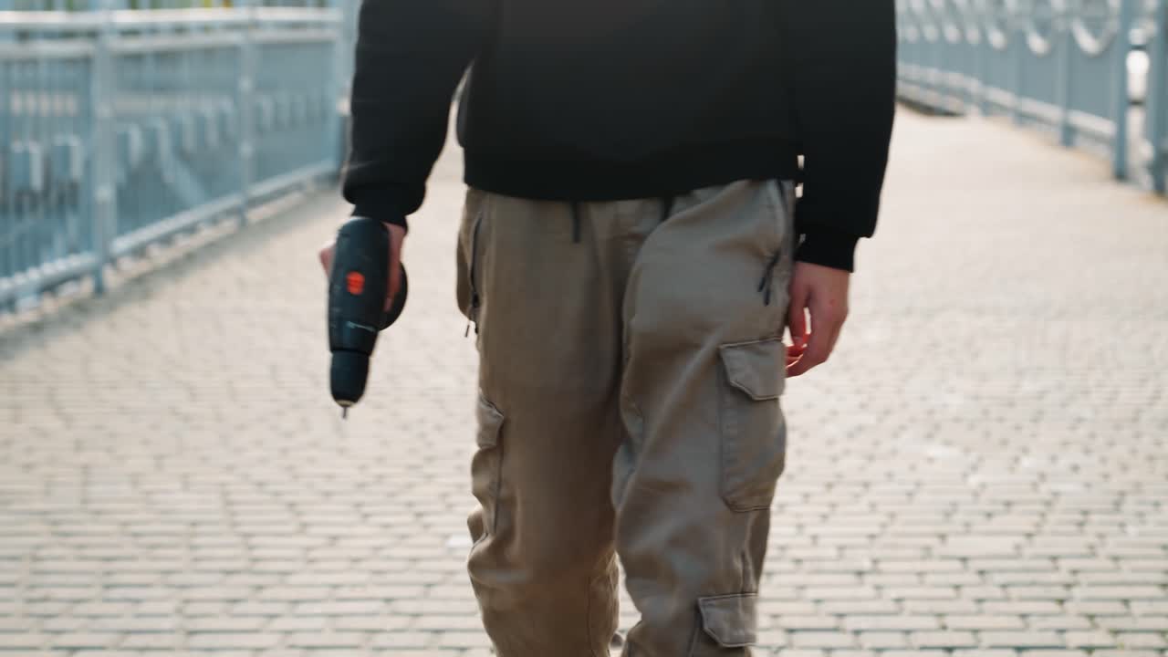 Caucasian man contractor walking across bridge holding power drill, cargo pants with tool pockets, determined stride, sunlit cobblestone path and utilitarian work atmosphere, closeup on tool