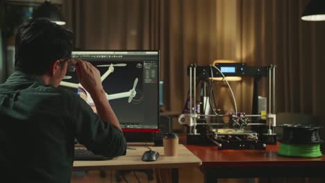 back view of frustrated asian engineer working on personal computer and 3d printer, screen shows cad software with 3d prototype of drone delivery