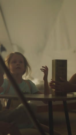 young mother tells son and daughter rules of board game while sitting on tourist chairs inside glamping site house. family evening of games and fun in nature