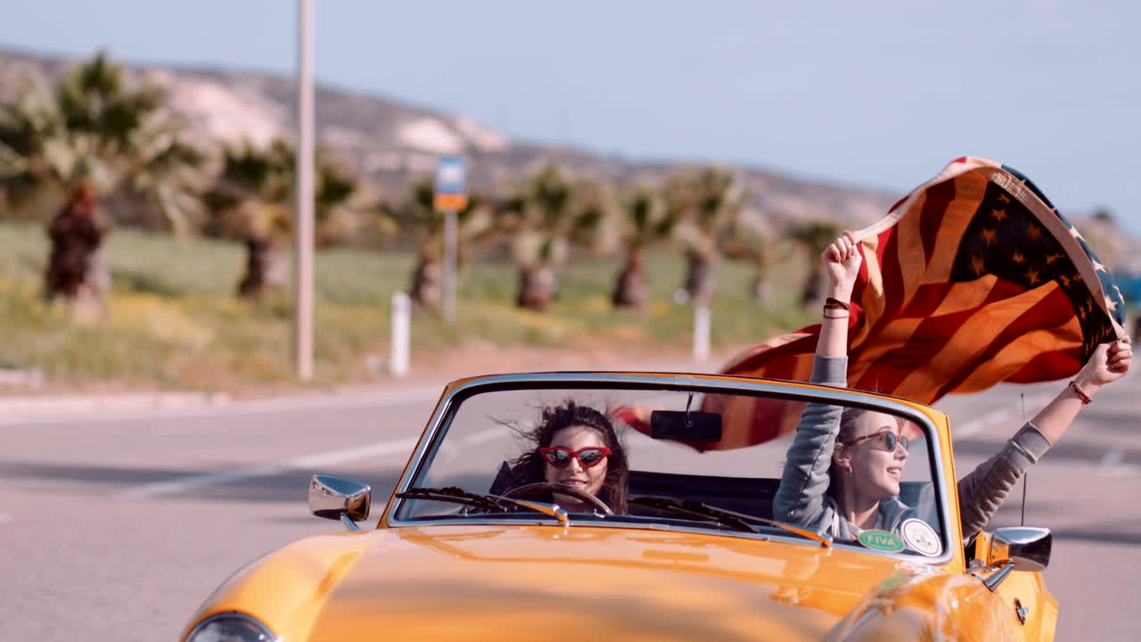 Junge Frauen in gelbem Retro-Cabrio feiern mit amerikanischer Flagge.