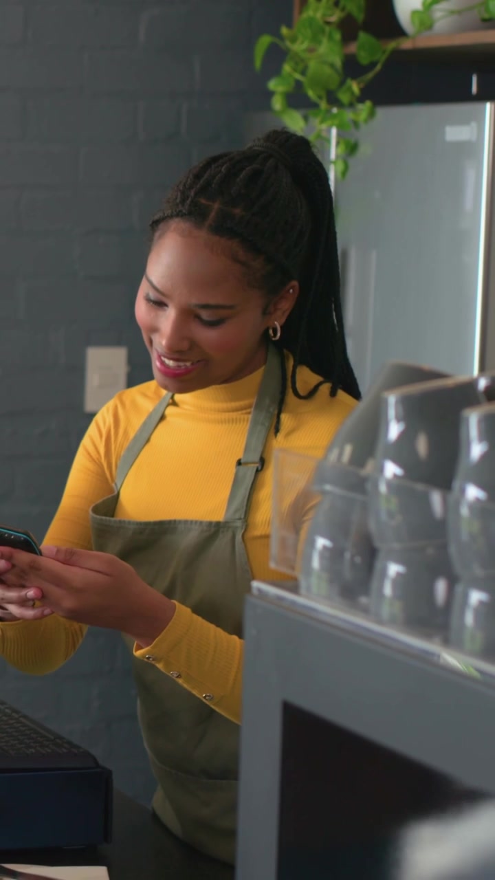 Vertical video: Taking order at cafe, barista in olive apron using POS and phone, handing cup
