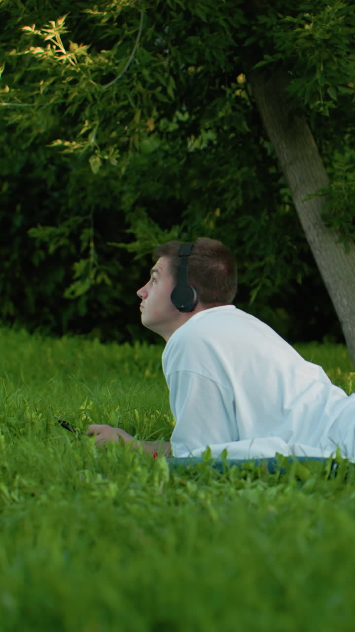 잔디에 누워있는 남자가 헤드폰, , 그리고 청바지를 입고, 음악에 머리를 흔들고, 울창한 초록색과 나무로 둘러싸여, 평온한 야외 순간을 즐기고 있습니다.