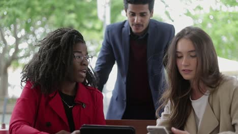 Two-women-and-man-in-summer-cafe-looking-at-gadgets,-discussing