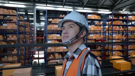 close up side view of asian male engineer with safety helmet standing in the warehouse with shelves full of delivery goods. looking around, checking the stock on racks