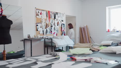 creative workspace filled with cut out fabric patterns on worktable, mannequin, design board, fabric rolls stacked by wall as two women finish discussion and leave room through door