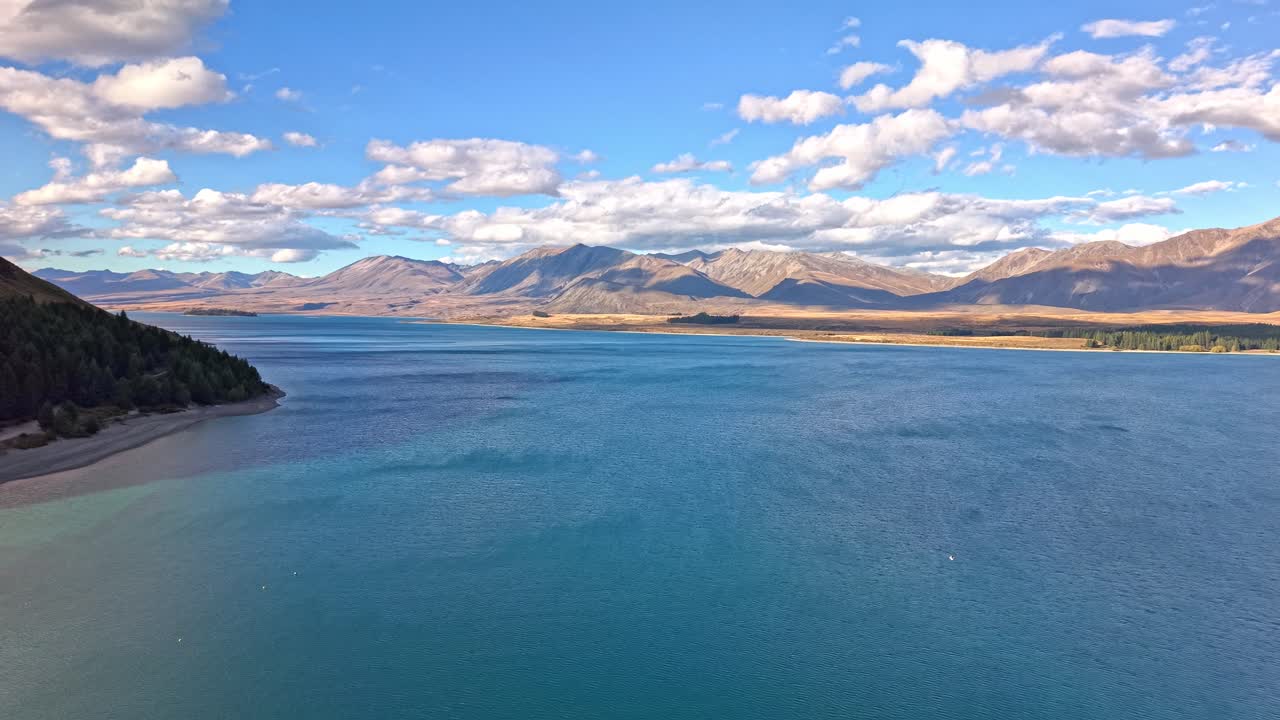 Aerial drone footage flying forward above Lake Tekapo, showing its blue alpine water, surrounding mountains, and the bright sky with clouds