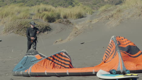 male in wetsuit checking rigging line as he walking back to kitesurf sail on windy beach in matanzas, chile