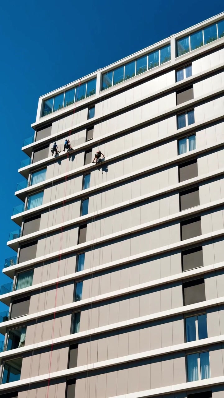Alpinistas industriais limpando a fachada de um edifício moderno sob céu azul claro. Três trabalhadores em cordas de segurança lavam janelas de vidro de um arranha-céu alto de concreto. Vista de baixo ângulo à luz do dia