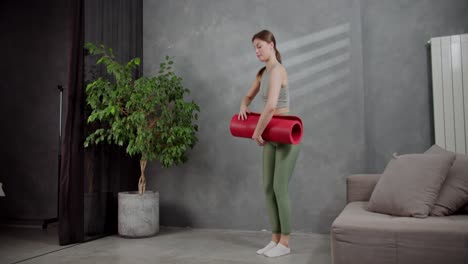 A-brunette-girl-in-a-gray-top-and-green-sweatpants-lays-out-a-red-mat-and-begins-her-exercise-in-a-modern-apartment-at-home.-A-young-brunette-girl-in-a-sports-summer-uniform-does-sports-on-the-red-carpet-near-a-houseplant-and-a-sofa-at-home