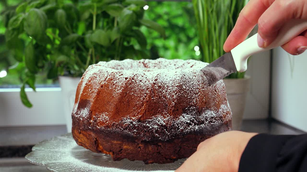 Static shot of a delicious marble cake while a woman's hand with a cake knife cuts off a piece of it for a delicious dessert from the kitchen with green plants in the background