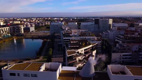 Vuelo-De-Drones-Sobre-Un-Lago-Con-Una-Fuente-En-El-Medio-De-La-Ciudad,-Rodeado-De-Vegetación,-Salones-Y-Edificios-Residenciales-Modernos-Con-Una-Chimenea-En-Funcionamiento