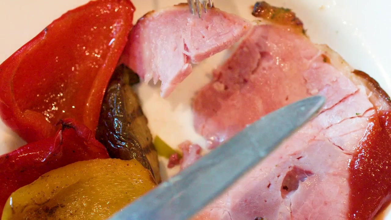 Primo piano di un coltello e una forchetta che tagliano del prosciutto arrosto servito con peperoni grigliati.