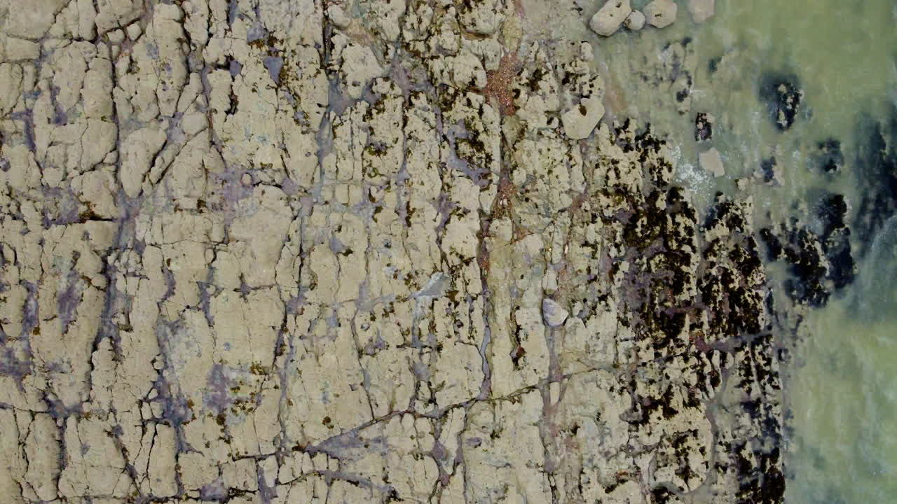 una vista aérea hacia abajo se desplaza sobre una costa rocosa de color marrón oliva, donde una tubería grande ingresa al agua de mar turquesa y se pueden ver efluentes en el agua circundante