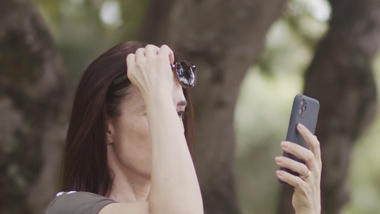 Mature woman stands in the park, phone in hand, sliding off sunglasses