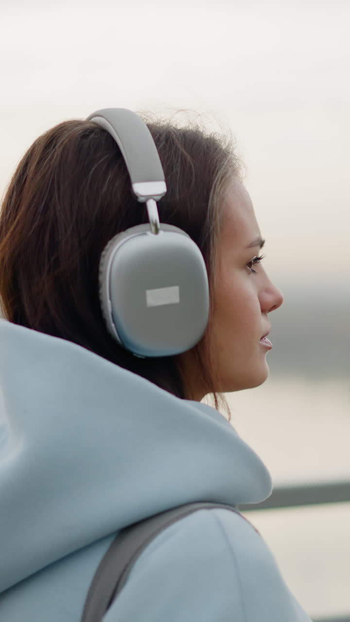 Close-up side view of young female student listening to music with headphones as she strolls near iron rail beside flowing water in peaceful outdoor setting, calm, relaxing walk by river