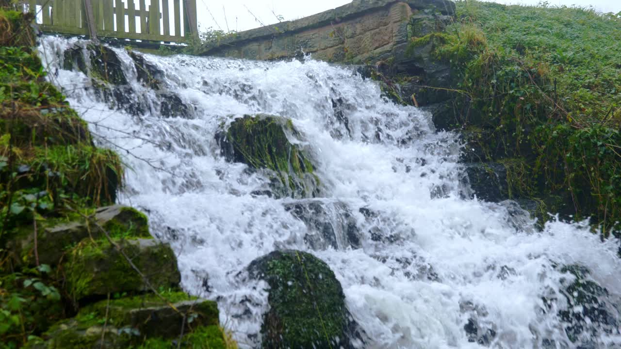 Close Up Of A Pond'S Waterfall Gushing Over Mossy Concrete Steps On A Foggy Winter Morning With Grassy Banks Either Side And A Wooden Bridge