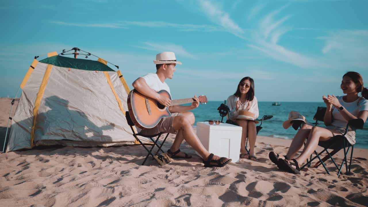 группа счастливых азиатских друзей играет на гитаре и поет с аплодисментами во время пикника и кемпинга на пляже на открытом воздухе летом.