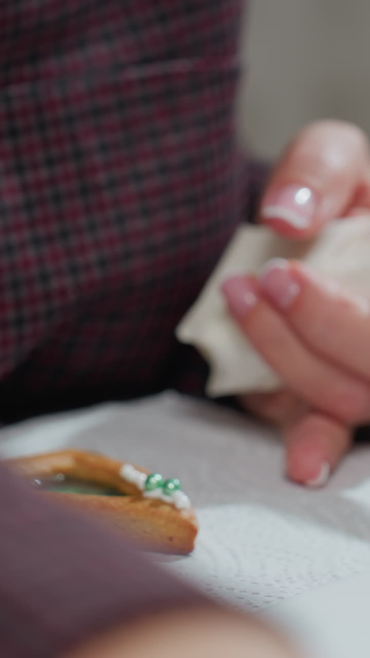 Close-up of baker holding white piping bag in one hand while using toothpick in other hand to adjust icing on cookie, focusing on careful decoration with precision on tissue paper surface
