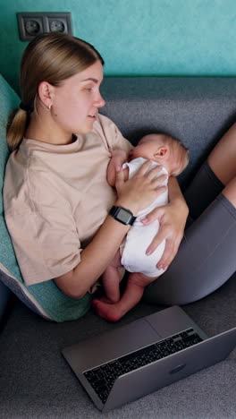 blonde caucasian woman with a newborn in hands sits on sofa. opened laptop is near the lady working from home. top view. vertical video