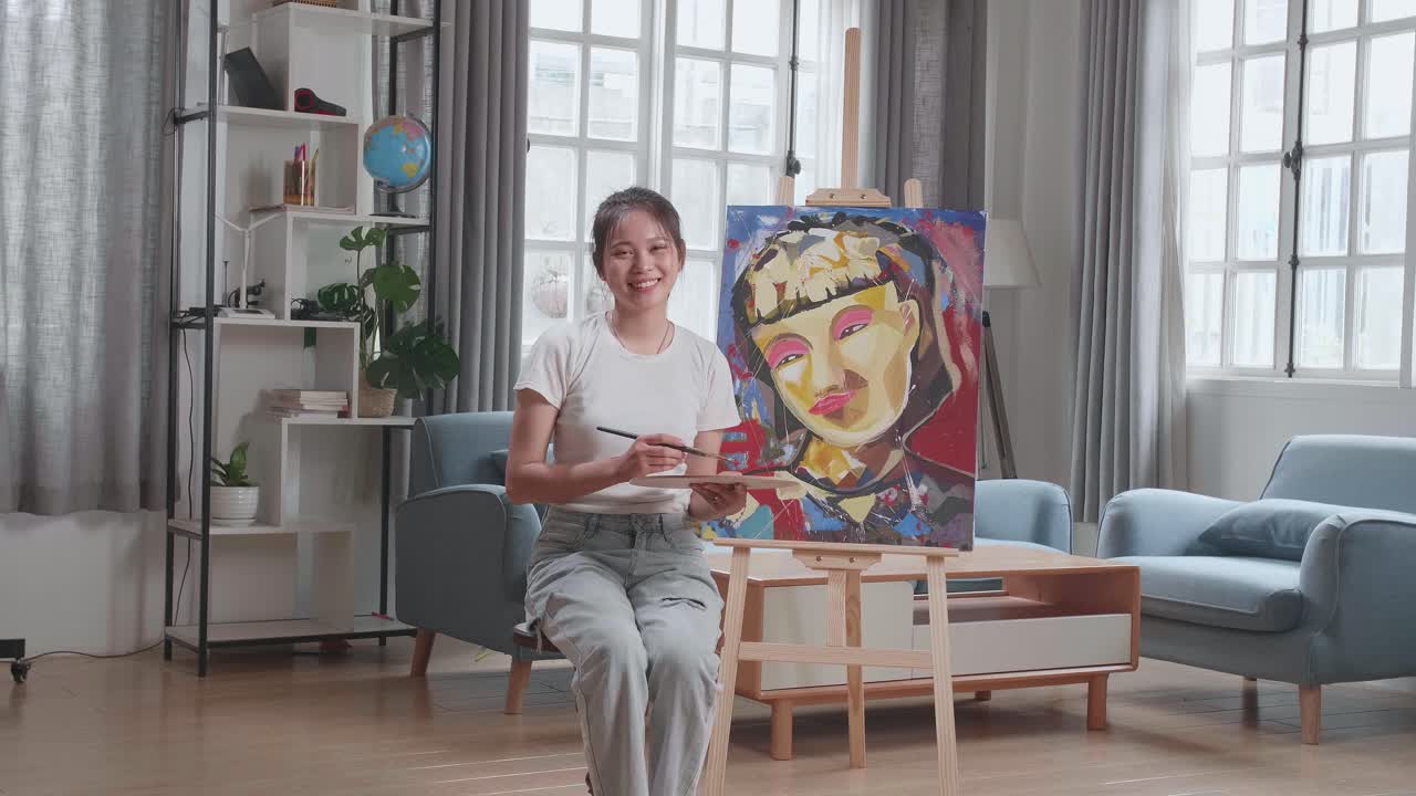 Asian Artist Girl Sitting On Round Wooden Chair Without Backrest Holding Paintbrush Mixed Colour And Smiling After Finish Painting A Girl On The Canvas