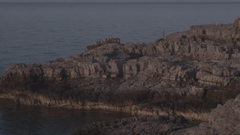 man with a hat fishing from a rock by beautiful sea, sunrise, golden hour, wide shot