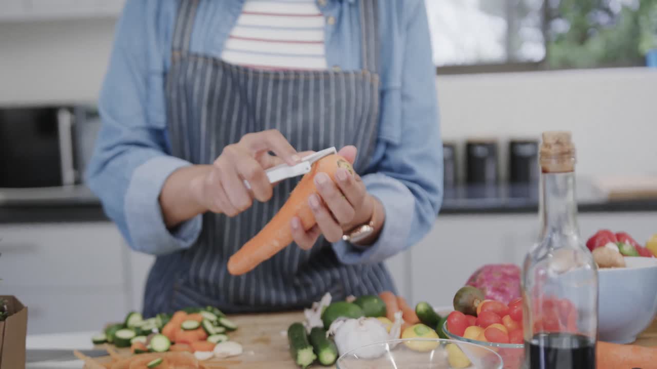 mujer biracial con delantal preparando comida, pelando zanahorias en la cocina, cámara lenta