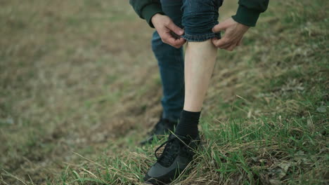 a close view of a man rolling down his jean trouser leg after inspecting his lower leg in an outdoor setting with a natural background of grass and earth