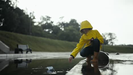 a teenage girl in a yellow jacket floats a paper boat through a puddle near a curb in a park