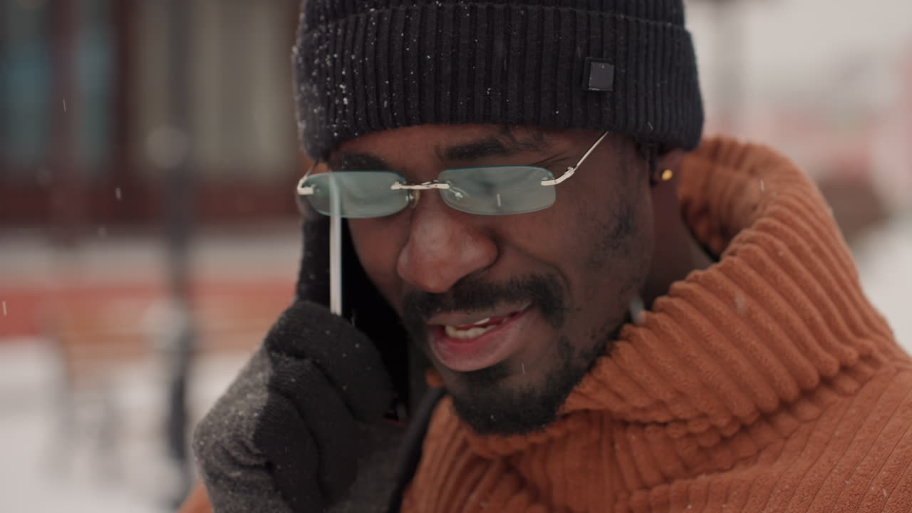Uomo di colore che sorride al telefono all'aperto, con berretto e maglione di lana, occhiali da sole e guanti, conversazione animata con risate, zaino in spalla, piazza cittadina con neve che cade, imprenditore fiducioso.