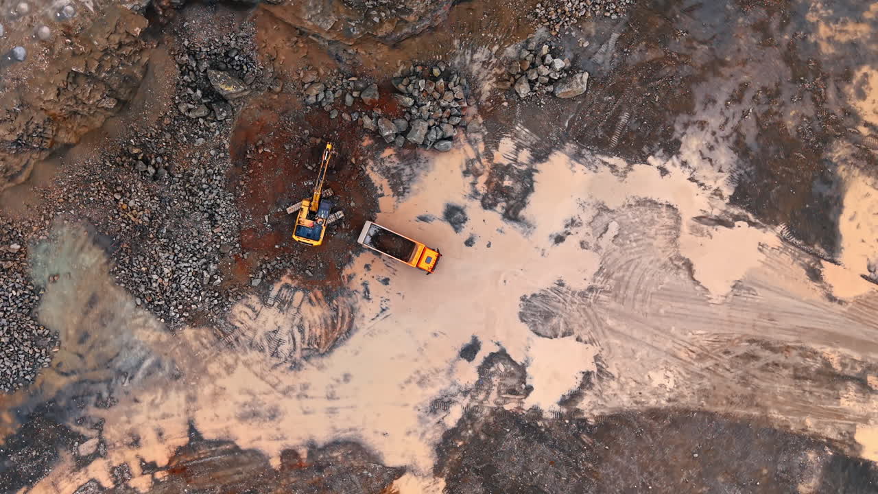 Top view of yellow excavator loading truck in quarry. Aerial shot of an excavator filling an orange dump truck with earth at a construction site