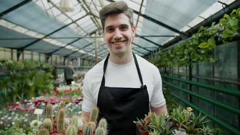 a young employee of a specialized store selling flowers holds flowers and cacti in their hands in pots and looks at the camera