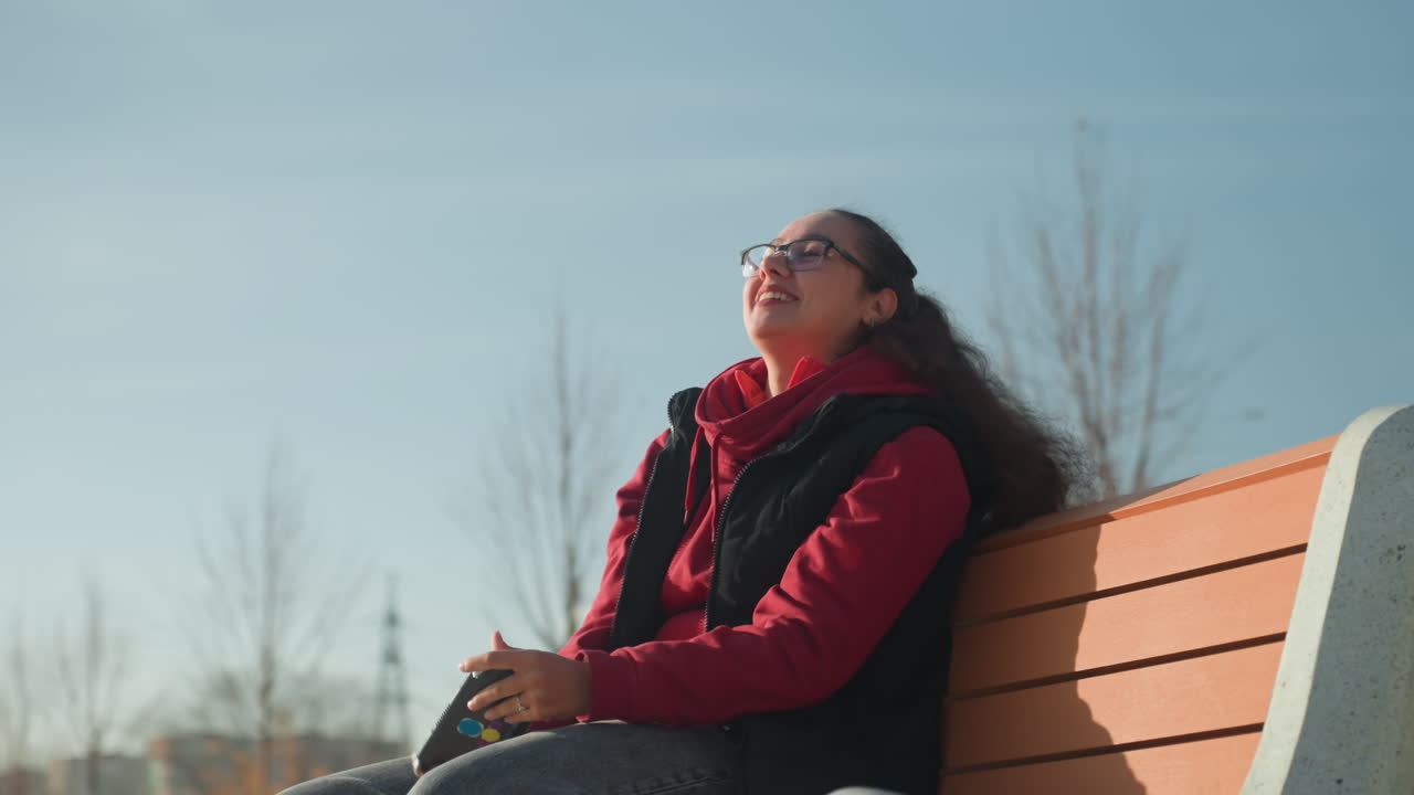 Femme sur un banc de parc prenant un selfie, étudiante détendue en pause capturant le ciel bleu avec son smartphone, lumière douce du soleil, veste d'hiver, lunettes, faible profondeur de champ, bâtiments urbains en arrière-plan, spontané.