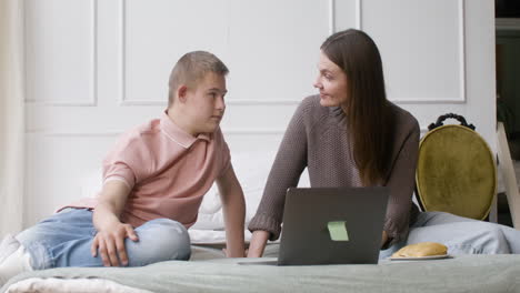 junge mit down-syndrom und seine mutter schauen etwas auf dem laptop zu hause im schlafzimmer auf dem bett