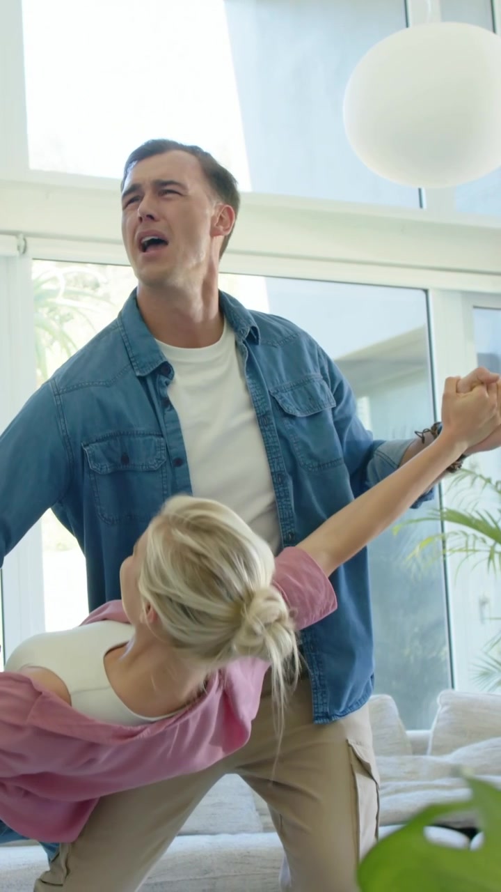 Vertical video: Reaching woman in pink lifting hand, man in denim guiding spin, dancing by sofa